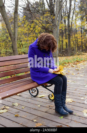 Lonely girl in the park on a bench with autumn leaves in her hands. The girl bowed her head. Stock Photo