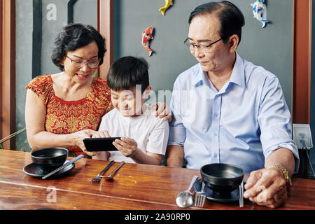 Happy Asian grandparents and grandchild sitting at cafe table and using mobile application on smartphone Stock Photo