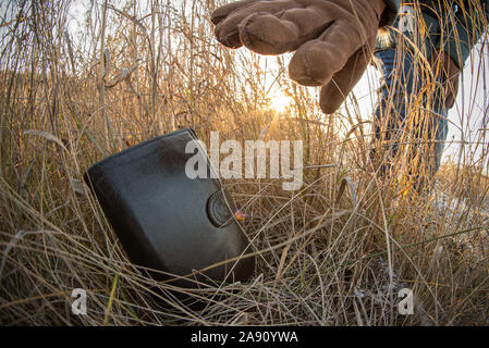Lost wallet in a grass on the ground outdoors and female hand picking up it from a ground. Close up. Fisheye view. Stock Photo