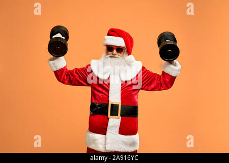 Happy mature Santa Claus with grey-haired beard in red sunglasses and christmas suit practicing with two heavy black dumbbells in studio. Physical exercise and healthy lifestyle concept. Stock Photo