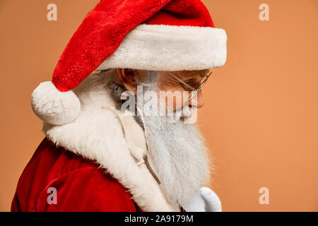 Side view of mature Santa Claus in red outfit and white gloves posing in studio with cute little mouse lying on white fur collar. Pretty rat is a symbol of 2020 new year. Stock Photo