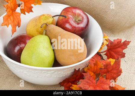 Fresh ripe pears and apples in a white bowl surrounded by autumn colored leaves. The background is a piece of burlap fabric. Stock Photo