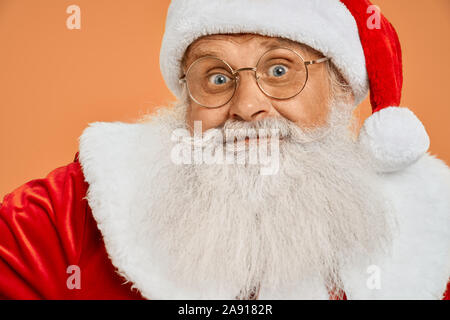 Funny senior bearded man in red Santa costume and eyeglasses looking straight to the camera with surprised facial expression. Santa Claus excited about christmas and new year Stock Photo