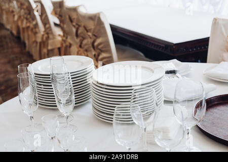 Assortment of kitchenware on white table.View over white round table with stacks of white plates, empty champagne flutes and glasses with bottles of Stock Photo