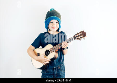 Portrait of a positive boy in casual clothes and hat playing ukulele. Copy space on white background. Stock Photo