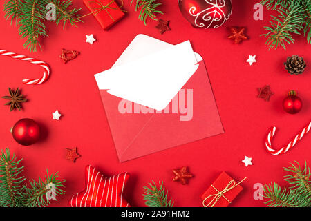 Christmas greeting card in frame made of fir tree branches, gift boxes, candy, red holiday decorations on red background. mock up. flat lay. top view Stock Photo