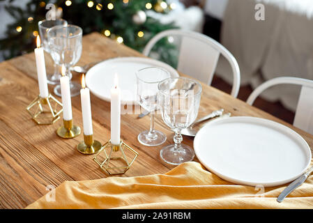 Table is served for a Christmas dinner in the living room, close-up view, table setting with a golden tablecloth, white plates, forks, knives, glasses Stock Photo