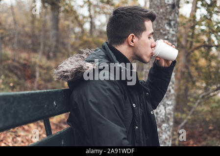 Young man sitted in a bench holding disposable coffee cup in the park in autumn season . Stock Photo