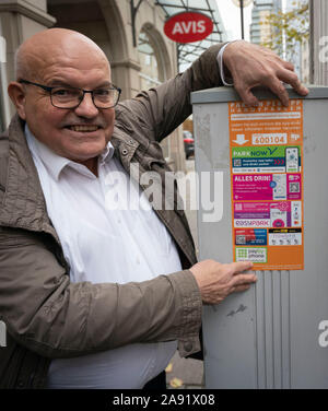 12 November 2019, Hessen, Frankfurt/Main: Traffic department Klaus Oesterling (SPD) sticks a sticker on the street in front of the traffic department with the providers of apps for the issuing of parking tickets via smartphone on a parking ticket machine, which is now also possible in Frankfurt am Main. In other Hessian communities, such as Wiesbaden, this has already been possible for some time. At the same time, prices for parking in the city centre are rising - four instead of three euros per hour are now due. Photo: Frank Rumpenhorst/dpa Stock Photo