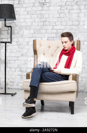 Teenager boy wearing red scarf and white sweater sitting in an armchair at home reading a book Stock Photo