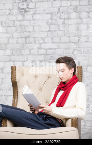 Teenager boy wearing red scarf and white sweater sitting in an armchair at home reading a book Stock Photo