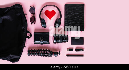 Trendy black accessories and contemporary gadgets on pink background, overhead view with copyspace. Modern headphones with heart symbol, camera lens, Stock Photo
