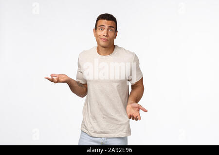 Clueless handsome young hispanic muscular man in casual t-shirt, shrugging, spread arms sideways, smirk and look indecisive, dont know how solve Stock Photo