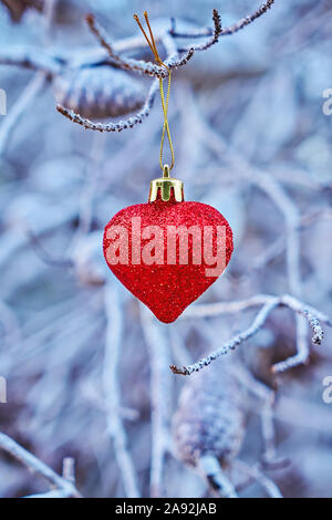 Christmas red glass heart-shaped ball on branches of tree. Vertical greeting card for celebration new year and Xmas. Selective focus, closeup Stock Photo
