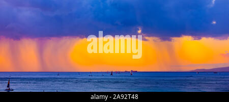 Storm clouds with rain and clouds glowing a bright yellow at sunset off Waikiki Beach with a silhouetted sailboats in the water Stock Photo