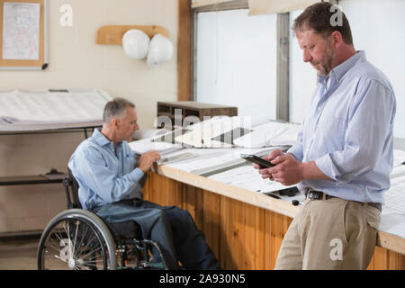 Two project engineers using their tablets to check job site plans, one in a wheelchair with a Spinal Cord Injury Stock Photo