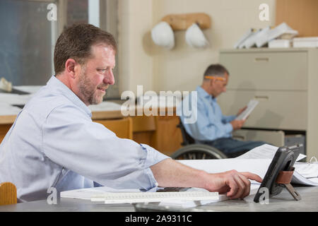 Two project engineers working at the job site office, one in a wheelchair with a Spinal Cord Injury Stock Photo