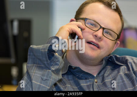 Hospital aid worker with Down Syndrome sitting at his desk and using his cell phone in office Stock Photo