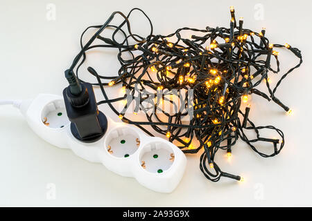 Checking shining christmas garland on a white surface. Black power plug of electric garland inserted into a white extension socket. Top view. Stock Photo
