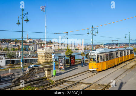 Budapest, Hungary - Nov 6, 2019: Yellow tram at public station. Danube river and the historical center with Fisherman's Bastion and Matthias Church in the background. People on the street. Sunny day. Stock Photo