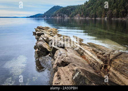 Layered, titled rock at the water's edge in BC's Gulf Islands points up a channel, with submerged shell middens revealing its First Nations history. Stock Photo