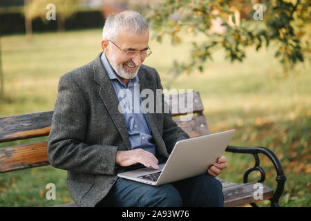 Senior businessman using laptop outside. Elderly man in gray jacket use laptop in the park Stock Photo