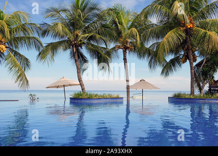 Luxury infinity swimming pool with blue water, umbrellas, palms and endless ocean view. Bali, Indonesia. Stock Photo