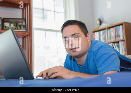 Man with Down Syndrome lying on bed using a laptop Stock Photo