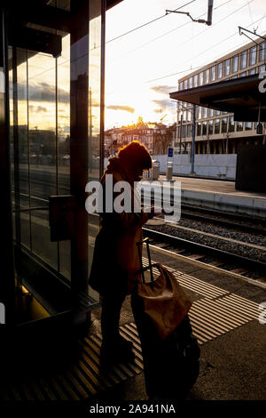 A woman stands with her suitcase on the platform of a train station beside the tracks and uses her smart phone; St. Gallen, St. Gallen, Switzerland Stock Photo