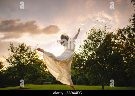 Dancer leaps against cloud-filled sky in park at sunset in gold light Stock Photo