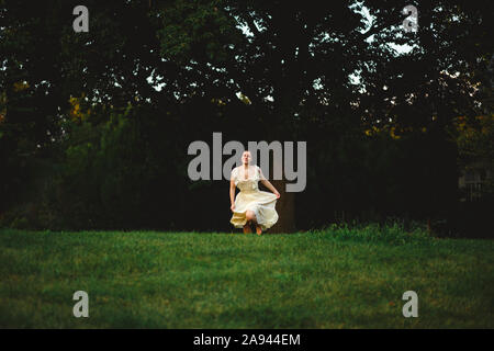 A young woman in a flowing dress runs across a darkened field by woods Stock Photo