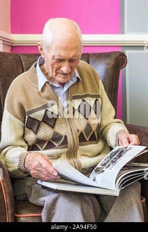 Senior man sitting in a chair looking at a book about World War 2; Hartlepool, County Durham, England Stock Photo