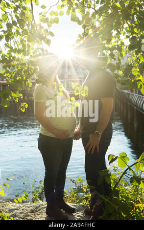 A mid adult couple during a pregnancy as they stand at the water's edge with a sunburst between them; Vancouver, British Columbia, Canada Stock Photo