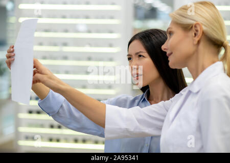 Standing near patient. Blonde-haired eye doctor wearing white coat standing near her patient reading the text Stock Photo