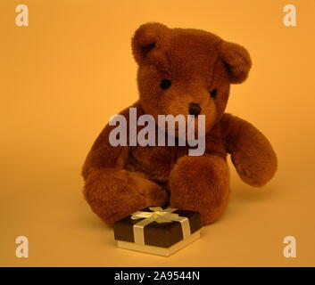 One cute brown teddy bear with small decorative gift box tied with golden bow close up, sits on pastel honey color background. Stock Photo