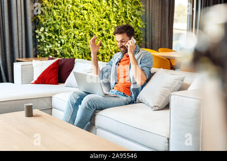 Leisure at Home. Young man sitting on sofa with laptop talking on phone laughing excited amazing news Stock Photo