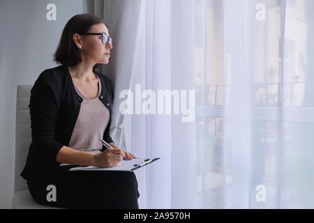 Serious business mature woman with clipboard sitting near the window Stock Photo