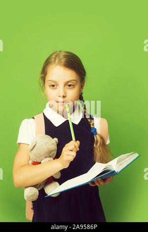 Kid in school uniform isolated on green background. Study and back to school concept. Girl with braids and thoughtful face expression. Pupil holds blue book, marker and teddy bear. Stock Photo