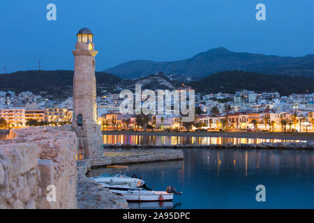 Rethymno, Crete, Greece. View along wall of the Venetian Harbour to illuminated 16th century Turkish lighthouse, dusk. Stock Photo