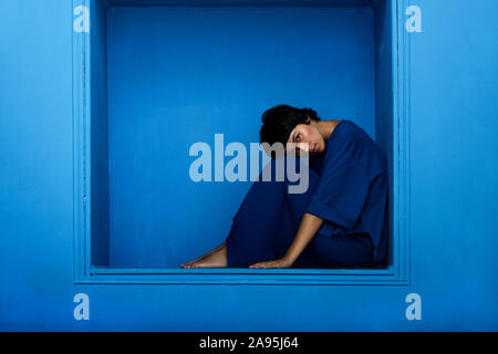 Young beautiful woman sitting in niche on blue background. Studio shot.