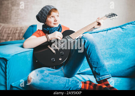 Attractive cute child in jeans, hat and red T-shirt dreamily plays with a smile on a black electric guitar sitting on the sofa at home Stock Photo