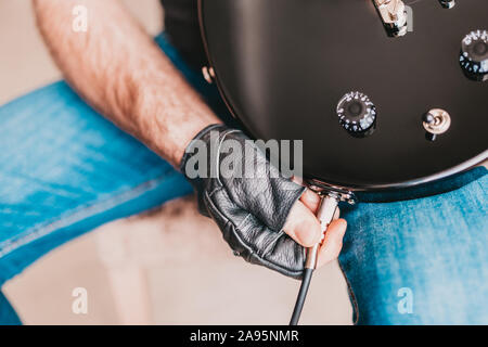 A man in black leather gloves connects a cable from a guitar amp combo to a black electric guitar Stock Photo