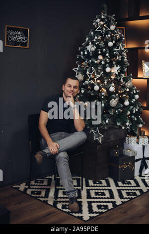 Handsome man against illuminated Christmas tree.fashionable watch looking away with hand on chin and pensive look. Stock Photo