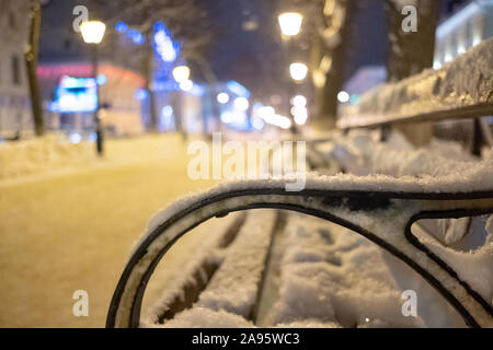 Fragment of a bench covered with snow on a night boulevard in the city in the light of lanterns. Winter cityscape in the defocused background. Stock Photo