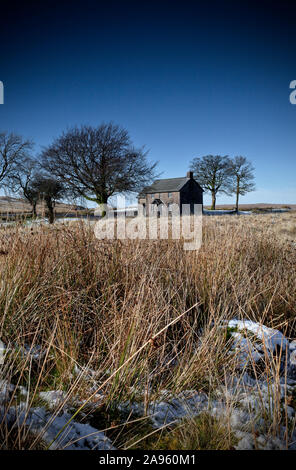 Remote solitary stone cottage surrounded by small group of trees in empty moorland landscape with light snow on ground and blue sky Stock Photo