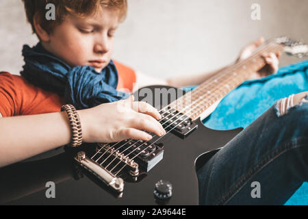 Informal red-haired teenager in jeans, hat and red t-shirt plays rock on a black electric guitar sitting on the sofa at home Stock Photo