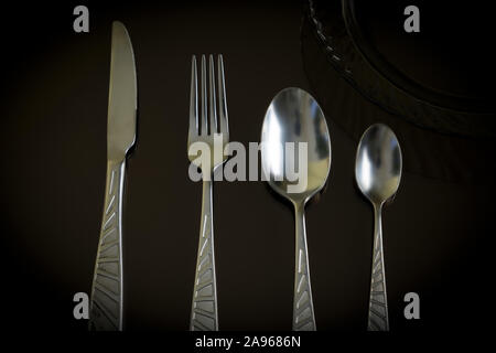 Still life of the kitchen objects. Dark glass plate, silver knife, fork, spoon, teaspoon on the dark reflective backdrop. Stock Photo