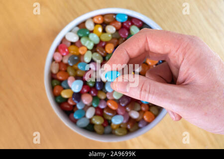 An adult man's hand holding a jellybean above a bowl filled with jellybeans. Concept - addiction to sweets, sweeties, candy Stock Photo