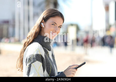 Happy girl holds mobile phone listening to music looking at camera standing in the street in winter Stock Photo