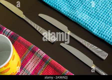 Blue and colored lines table-napkins and cutlery as a still life on dark reflective table. Three silver knives between blue and multicolor napkins. Stock Photo